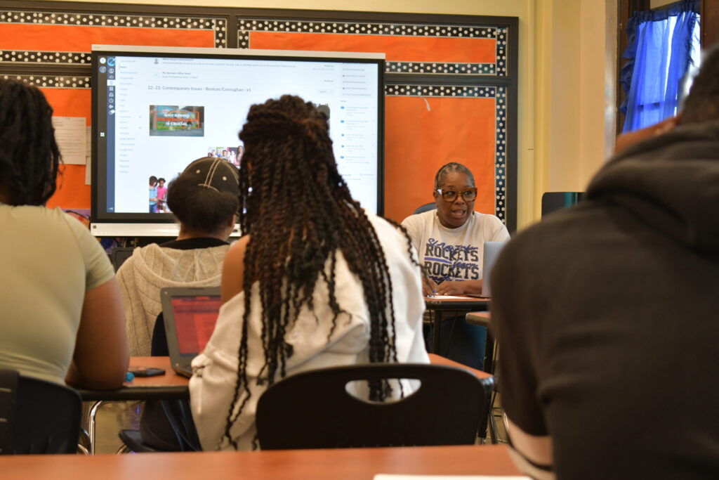 A Black teacher sits in a classroom with a screen behind her and students looking at her.