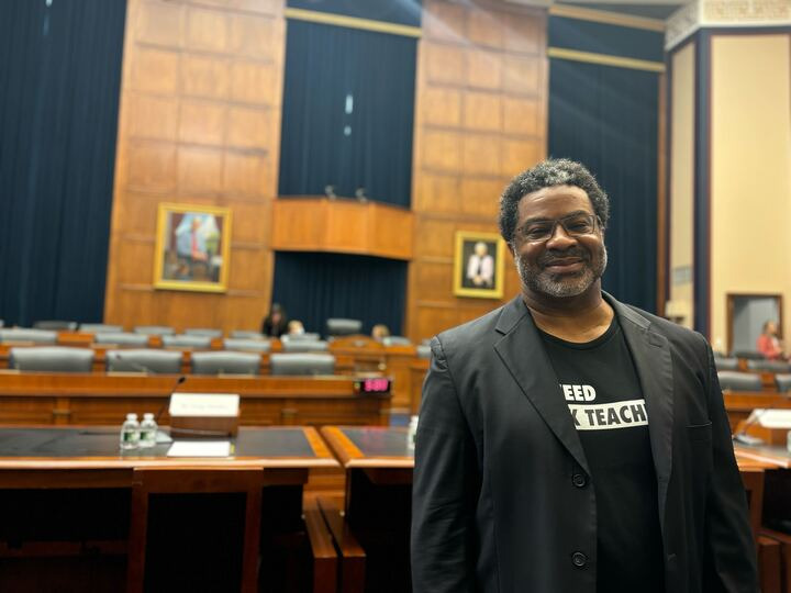 A man smiles inside an official hearing room wearing a blazer with a We Need Black Teachers shirt.